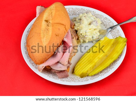 Cold cut hoagie sandwich with ham, roast beef and peppered turkey breast on roll and served with potato salad and bread and butter pickles against red background.
