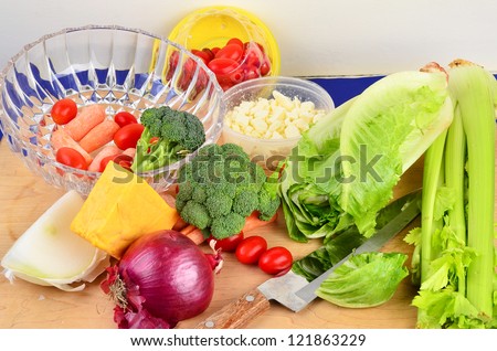 Various ingredients for making a salad on cutting board with knife and clear crystal bowl.  Included are onions; cheese; tomatoes; carrots; lettuce; celery; broccoli.