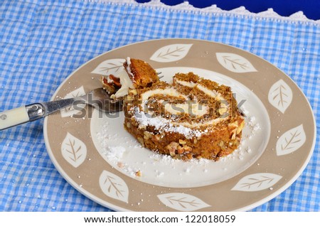 Bite of pumpkin role on fork being cut from large slice on plate with blue gingham place mat as background.  Realism added by sprinkles of confectioners powdered sugar on plate and place mat.