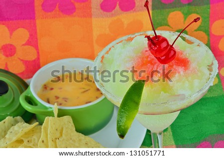 Frozen margarita with maraschino cherry and lime on brightly colored background served with chili con queso dip and tortilla chips.