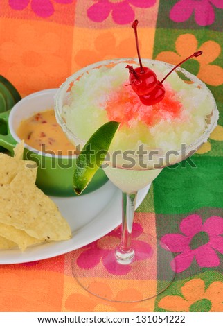 Frozen margarita with maraschino cherry and lime on brightly colored background served with chili con queso dip and tortilla chips.