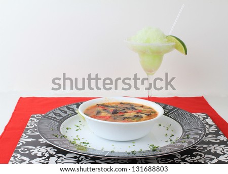 Frozen lime margarita with bowl of tortilla soup in black, red and white color scheme with Mexican Aztec graphics on platter.