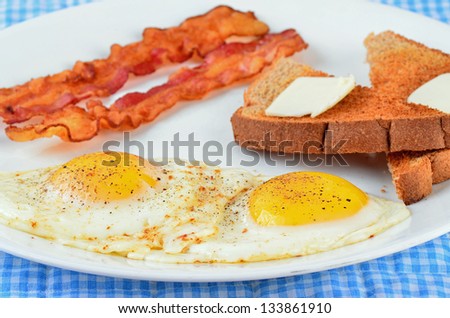 Fried eggs sunny side up on white plate with bacon and toast with butter in blue gingham setting.