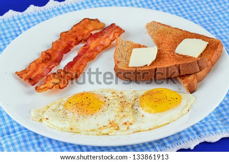 Bacon with eggs sunny side up on white plate against blue and white background with toast and butter.