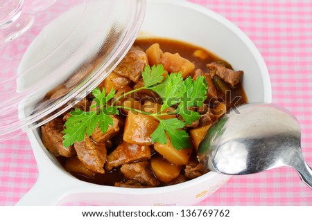 Steaming hot beef stew in serving bowl with spoon on pink gingham background.