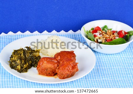 Country Cooking - Meatballs in tomato sauce on white plate with mashed potatoes and collard greens against blue gingham place mat.  Served with tossed green salad.