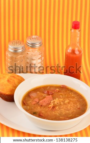 Spicy Cajun Cooking - Sausage Gumbo in white bowl on white plate with cornbread muffin against colorful orange and yellow background.  Salt and Pepper Shaker and bottle of Louisiana Hot Sauce.
