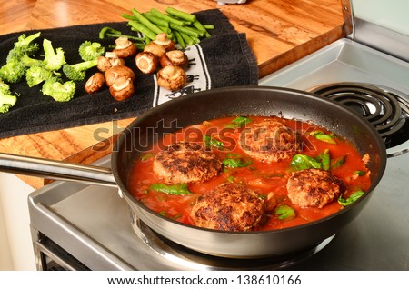 Hamburger steaks in tomato gravy simmering on hot electric stove with vegetables being prepared on wooden counter top.
