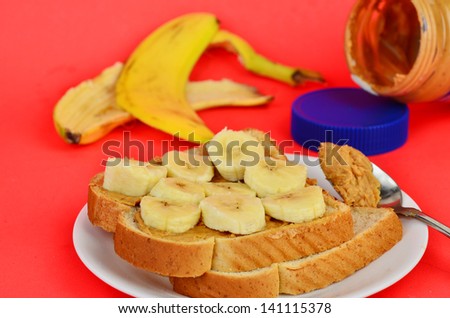 Crunchy peanut butter on honey wheat bread with banana bites.  Peanut butter jar and banana peel on red background.
