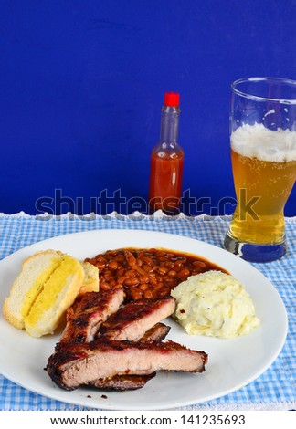BBQ Ribs on white plate with potato salad and baked beans.  Blue background with bottle of hot sauce and glass of cold beer.