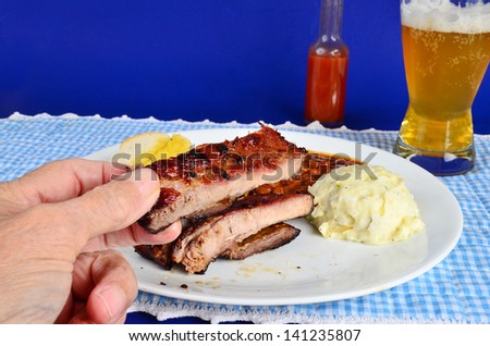 Man\'s hand holding charbroiled rib with baked beans and potato salad on white plate in background.  Served with beer and buttered garlic bread against blue background.