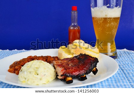 BBQ Spare Rib Dinner with glass of beer and bottle of hot sauce on blue background.  Plate includes baked beans, potato salad, and buttered garlic bread.