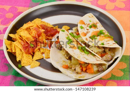 Mexican Burrito - Meat and potatoes with chopped vegetables on flour tortilla.  Garnished with diced jalapeno and spicy sauce; served with chips and salsa on brightly colored floral pattern place mat.