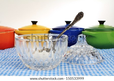 Elegant antique cut glass crystal sugar bowl with lid and sterling silver sugar spoon on blue gingham place mat with colorful containers in background.