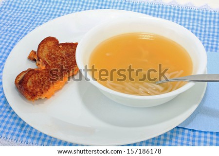 Half a grilled pimento cheese sandwich and bowl of chicken noodle soup on blue gingham place mat.