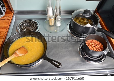Cooking soul food with creamed corn in cast iron skillet; black eyed peas in small sauce pan and collard greens in medium sauce pan with lid -- all simmering on stainless steel electric range.
