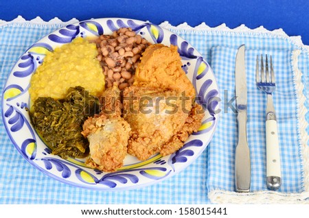 Country fried chicken dinner with collard greens; creamed corn; black eyed peas; and candied yams on blue plate against blue gingham.