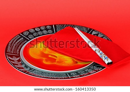 Colorful place setting in oriental restaurant with bright red plate in black charger plate with geometric design and white chopsticks on red napkin.