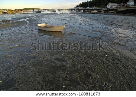 Stranded white dinghy sits stranded on mud flats of low tide with Stonington Harbor Maine fishing fleet in background.  Soft evening light. Copy space at bottom.