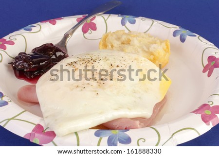 Fried egg and ham on biscuit with grape jam.  Served on paper plate against blue background.