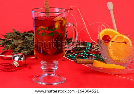Clear glass coffee mug with holiday design filled with mulled wine -- cherry; orange slice; and cinnamon stick.  Mug surrounded by Christmas decoration.