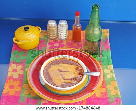 Bowl of chili covered with cheese strips on brightly colored Mexican Restaurant setting with bottle of beer.