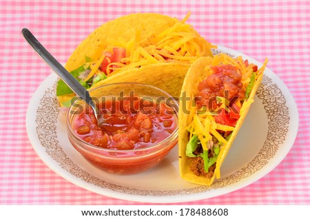 Clear bowl of spicy salsa on plate with two crunchy tacos on corn tortillas.