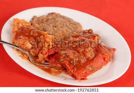 Mexican dinner with beef enchiladas in red salsa (Enchiladas Rojo) with rice and beans on white plate against red background.