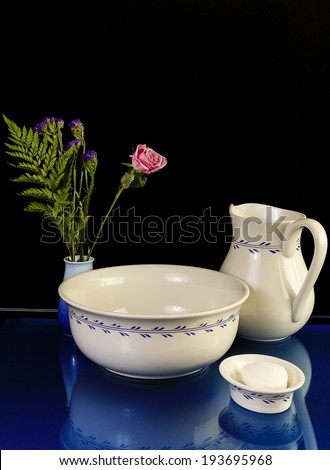 Still Life with antique wash basin with matching water pitcher and soap dish reflecting in glass-top table with flowers in vase.