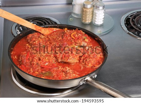 Skillet filled with chicken thighs covered in spicy tomato sauce and paella ingredients cooking on electric range.