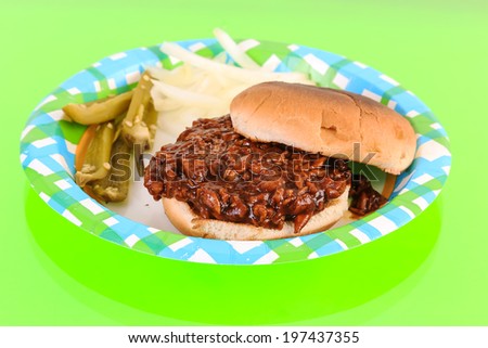 Pork Barbecue Sandwich on Paper Plate with onions and jalapeno peppers reflecting in green glass-top counter.