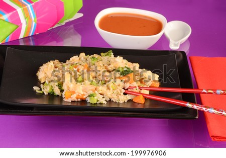 Chicken and rice stirfry on black rectangular plate against purple reflective surface with red chopsticks.  Bowl of soup with soup spoon and carry out cartons in background.