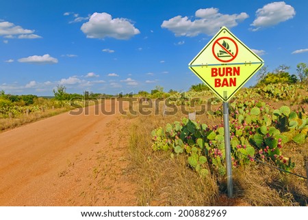 Burn Ban sign on gravel road through dry grass field and cactus emphasizes seriousness of drought in Central Texas.