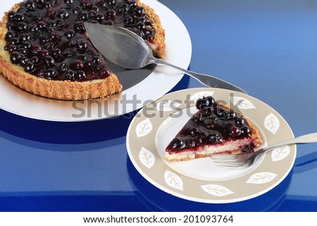 Serving of blueberry cheesecake on dessert plate in foreground with rest of cake on platter in background.  Reflection in blue glass table top.