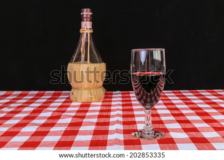 Glass of Chianti wine on red and white checked table cloth with basket bottle against black background.