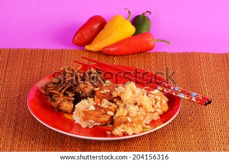 Brightly colored setting with sweet and sour chicken dinner on red plate with General Tso\'s Chicken against woven bamboo place mat and assorted peppers.