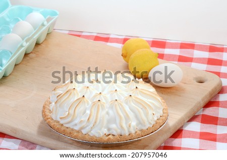Lemon Meringue Pie in country kitchen setting on wooden cutting board with some ingredients against red plaid tablecloth background.