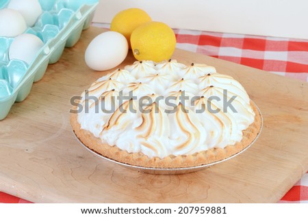 Lemon Meringue Pie in country kitchen setting on wooden cutting board with some ingredients against red plaid tablecloth background.