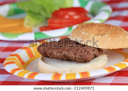 Closeup of hamburger patty on sesame bun on paper plate with vegetable and cheese ingredients in background on red plaid tablecloth.  Selective focus with shallow depth of field.