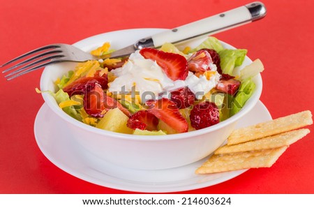 Chunks of pineapple and strawberries over bed of romaine lettuce and cottage cheese with crackers on red background.