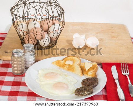 Organic Sunny Side Up eggs on white plate with sausage patties in Country Setting with wire basket of fresh farm eggs in background.  Shells from cracked eggs on wooden cutting board.
