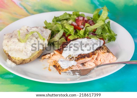Taking bite from salmon steak on white plate with stuffed baked potato and garden salad; background is simulated ocean scene.
