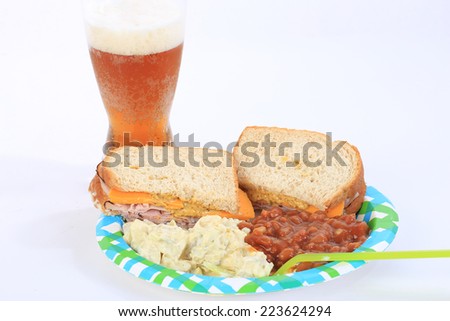 Ham and Cheese on honey wheat bread on paper plate with potato salad and baked beans with glass of beer.
