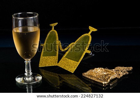 Drink glass, party mask and ladies evening purse on black reflective background.