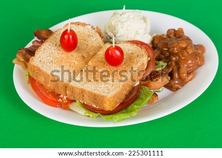 Bacon Lettuce Tomato Sandwich with baked beans and potato salad on white plate against green background.