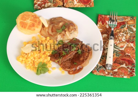Meatloaf dinner on white plate with corn and mashed potatoes and gravy against green background.