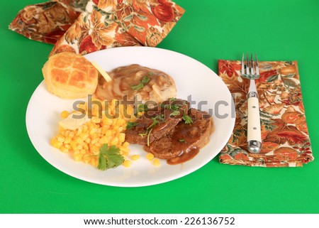 Meatloaf dinner on white plate with corn and mashed potatoes and brown gravy against green background.