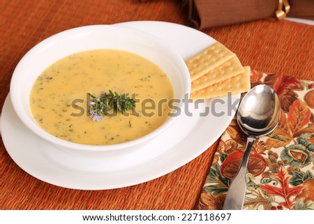 Autumn colors surround steaming hot bowl of cheese and broccoli soup garnished with sprig of rosemary with tiny purple bloom.