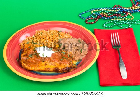 Dollop of sour cream on chicken enchiladas in red sauce in brightly colored plate with beans and rice.  Colorful green background with party beads.  Cheerful colors of festive celebration.