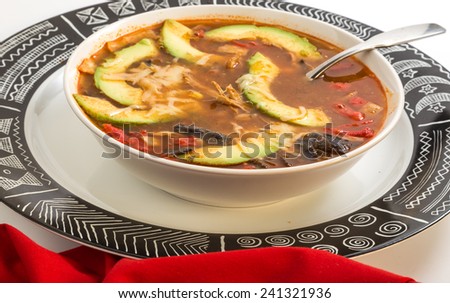 Tight shot of bowl of tortilla soup in white bowl on graphic black and white charger plate with red napkin. Ancient Aztec Pattern on plate.